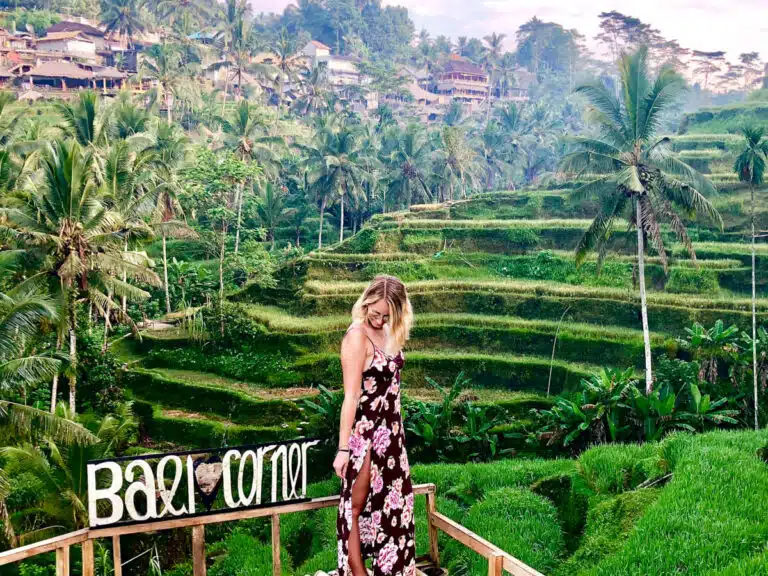 A woman in a floral dress stands on the bamboo platform labeled “Bali Corner,” smiling with layers of Tegallalang’s bright green terraces stretching behind her.