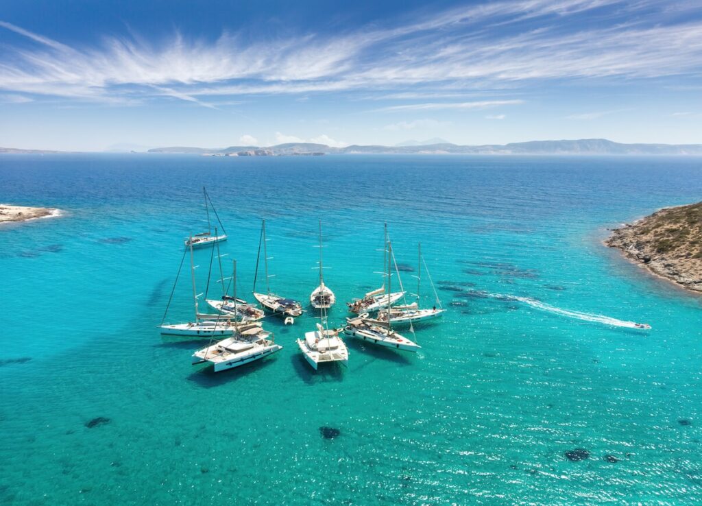 ailboats anchored in vivid turquoise water near Polyaigos island in Greece. The aerial view shows crystal clear sea surrounded by rocky coastline and distant islands.