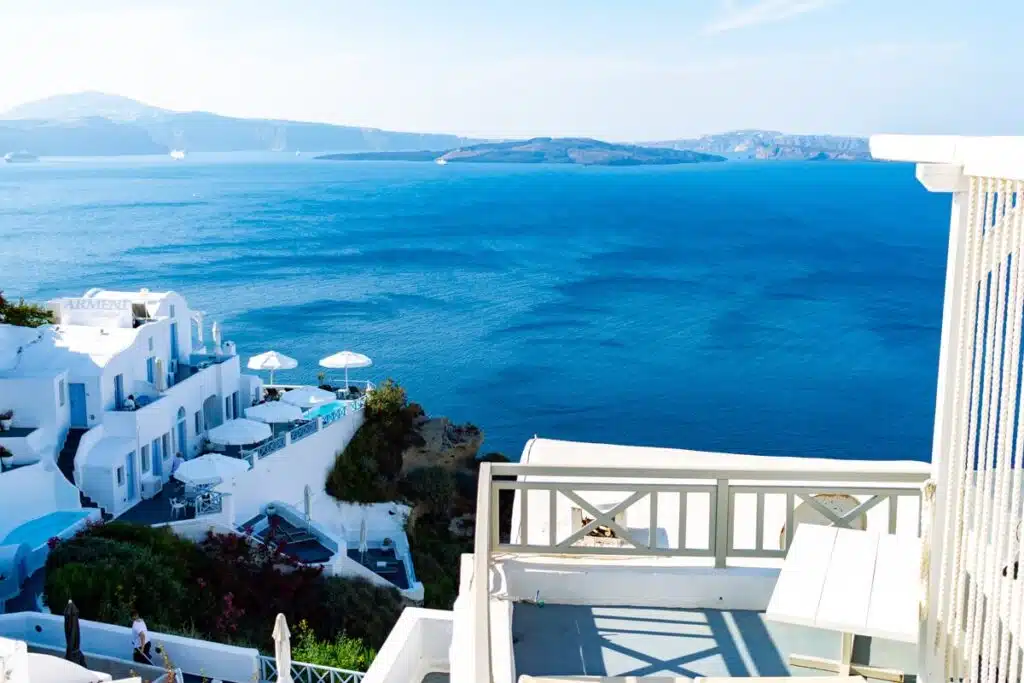 View from a balcony at Kaleidoscope Cave Houses in Oia, Santorini overlooking whitewashed cliffside buildings and the deep blue caldera. Patio umbrellas and terraces sit above the water with rocky islands visible in the distance under a clear sky.