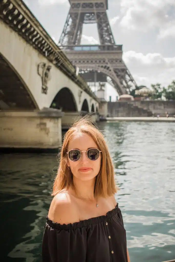 girl in black shirt and sunglasses standing underneath a bridge with the Eiffel Tower in the background in Paris