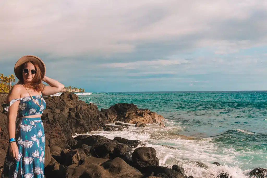 girl in tropical blue dress and hat standing on lava rocks on Big Island of Hawaii with ocean in background