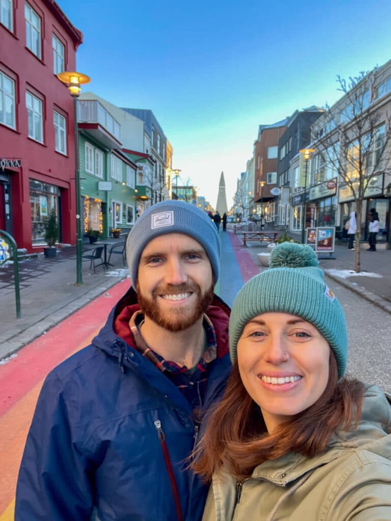 A smiling couple dressed in winter clothing takes a selfie on Rainbow Street in Reykjavik, Iceland in winter, with colorful buildings and the Hallgrímskirkja church in the distance.