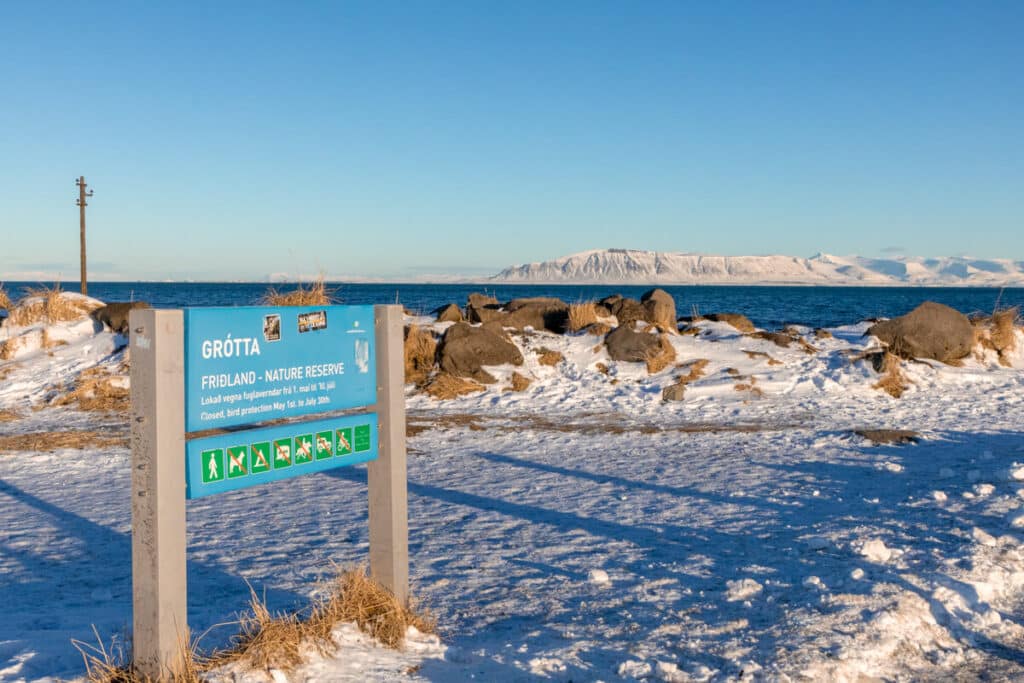 blue sign for Grótta Nature Reserve stands in the foreground, surrounded by snow and coastal rocks, with a clear view of the ocean and snow-covered mountains under a bright blue sky. The sign mentions seasonal closure for bird protection from May 1st to July 30th.