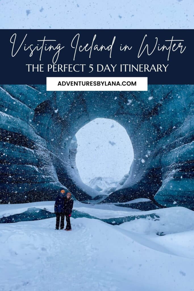 A couple stands in a dramatic blue ice cave while snow falls heavily around them, with bold script reading “Visiting Iceland in Winter: The Perfect 5 Day Itinerary” and the website adventuresbylana.com, ideal for Pinterest sharing of an Iceland winter itinerary.