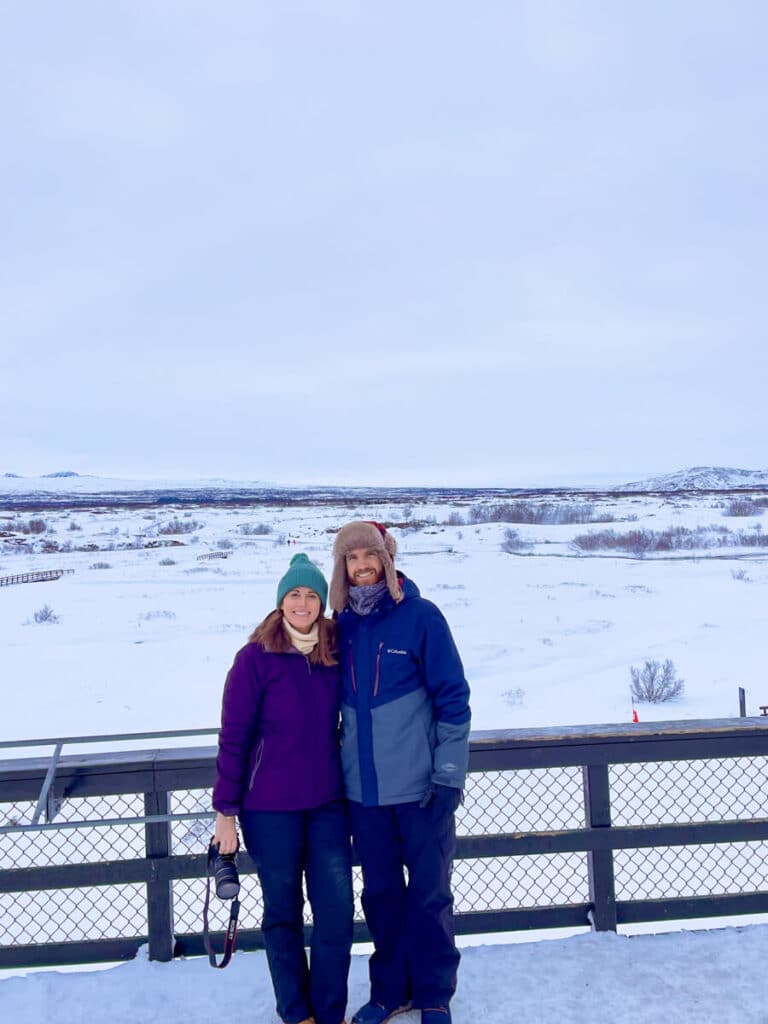 A couple in snow gear stands at a viewpoint overlooking a vast, snow-covered landscape in Thingvellir National Park, Iceland in winter, with camera in hand and mountains in the distance.