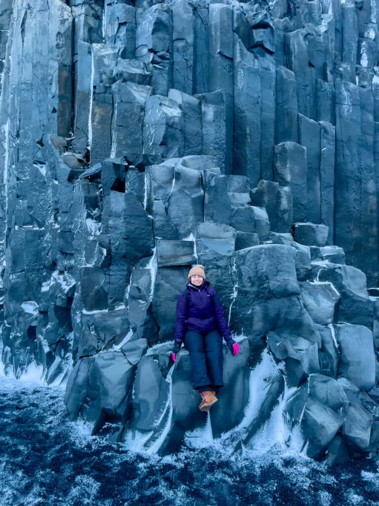 A woman bundled in winter clothing sits on striking black basalt columns lightly dusted with snow at Reynisfjara Beach in Iceland in winter, surrounded by dramatic volcanic rock formations.