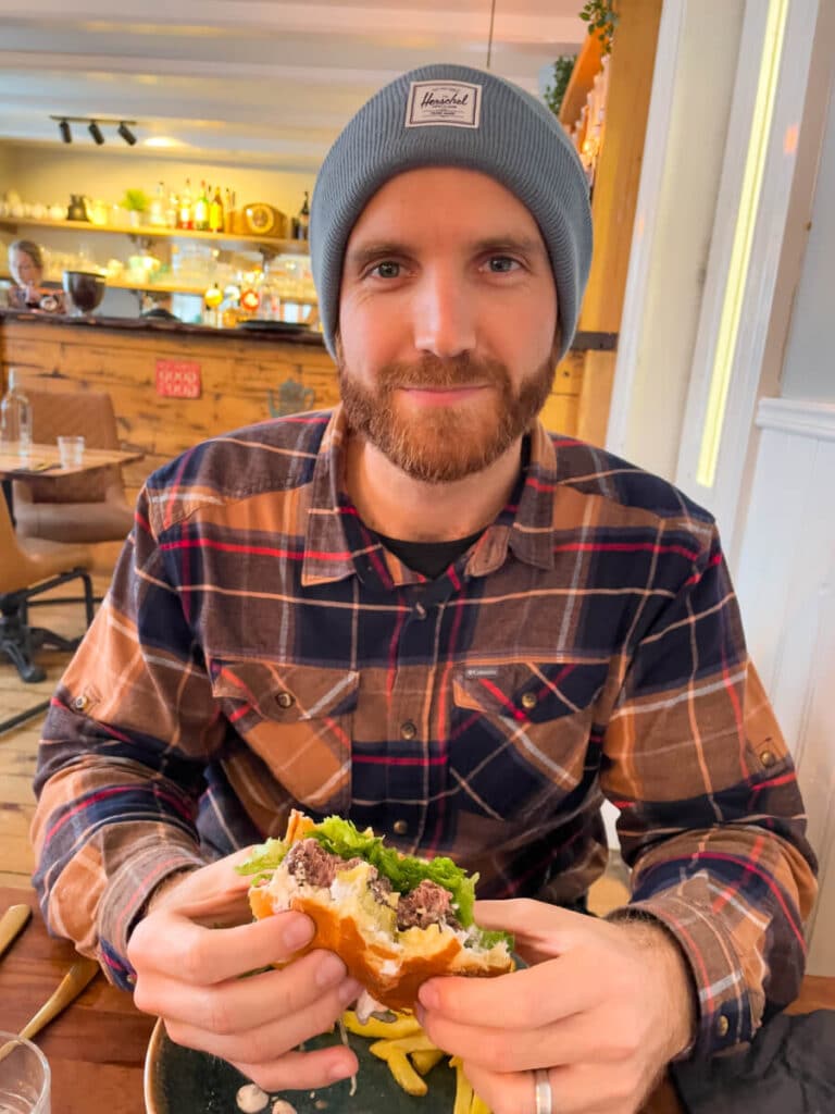 A man in a flannel shirt and beanie smiles while holding a half-eaten reindeer burger with fries on the table in front of him, enjoying a warm meal indoors during winter in Iceland.