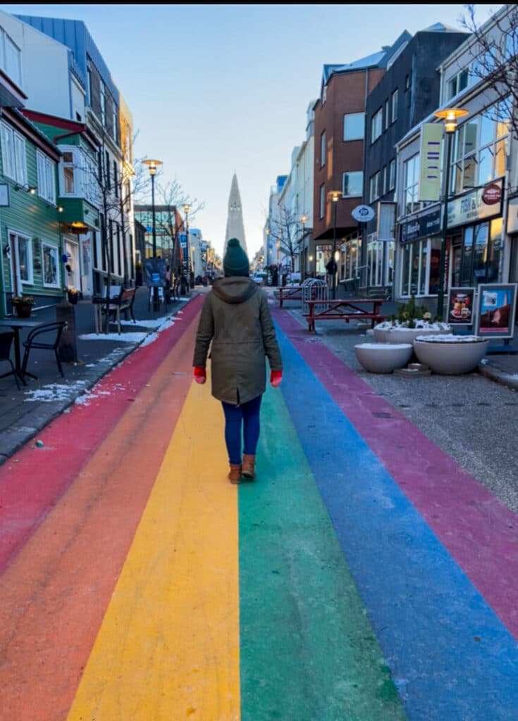 A woman in a green beanie walks along Rainbow Street in Reykjavik, Iceland in winter, heading toward the Hallgrímskirkja church under a clear blue sky.