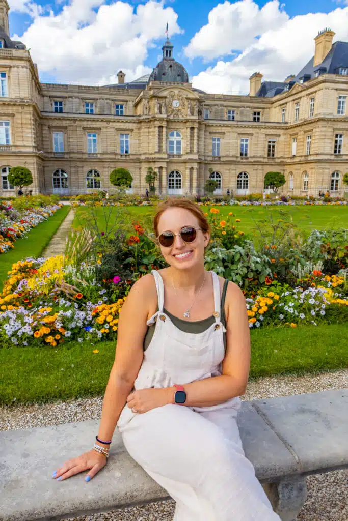 girl in beige jumpsuit sitting on wall in front of Luxembourg Gardens in Paris smiling at camera with beautiful flowers behind her