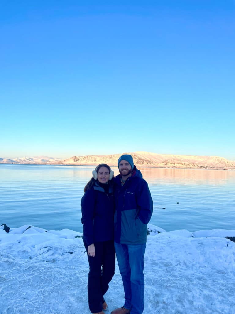 couple bundled in winter gear standing in the snow in Reykjavik, with the ocean and sunny mountain the background