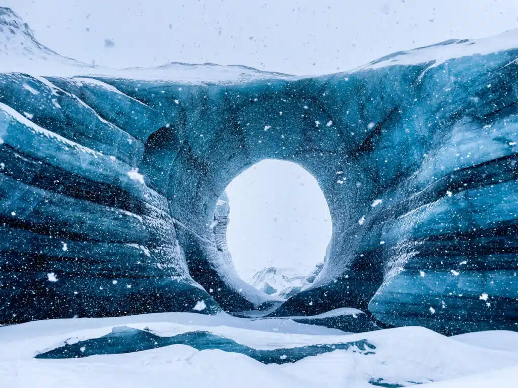 Snow falls gently around the entrance of the giant arch outside the Katla Ice Cave in Iceland, framed by massive blue-tinted glacial walls with a naturally formed circular opening in the center. The icy scene is dramatic and otherworldly, emphasizing the cave’s unique shape and scale.