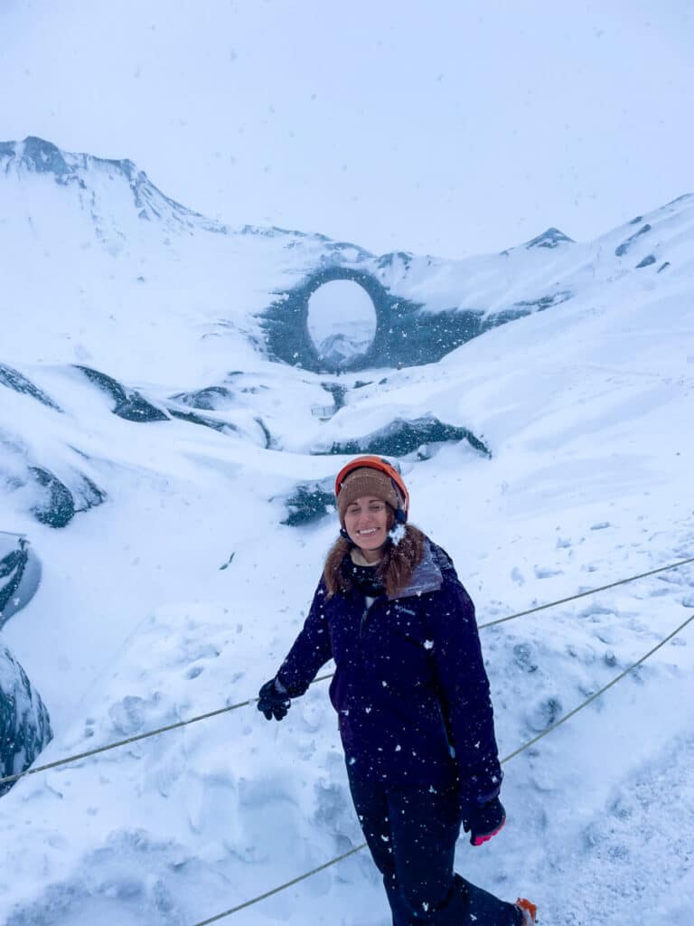 girl smiling at the camera with eyes closed in front of snowy landscape and large ice arch at the Katla Ice Cave in Iceland