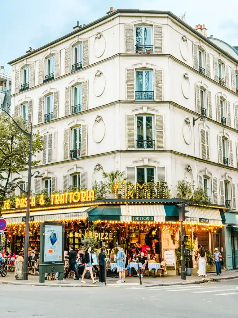 Corner view of the charming trattoria "Firmine", with striped green awnings, glowing string lights, and sidewalk tables filled with diners. The white building features classic French shutters and ornate molding, adding to the inviting neighborhood atmosphere.