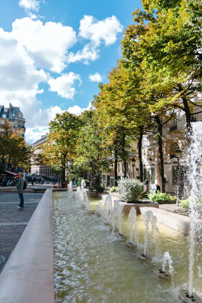 A row of small fountains splashing into a long rectangular basin surrounded by greenery and tree-lined streets in front of the Sorbonne in the Latin Quarter of Paris. A few pedestrians stroll nearby under a sky dotted with fluffy clouds.