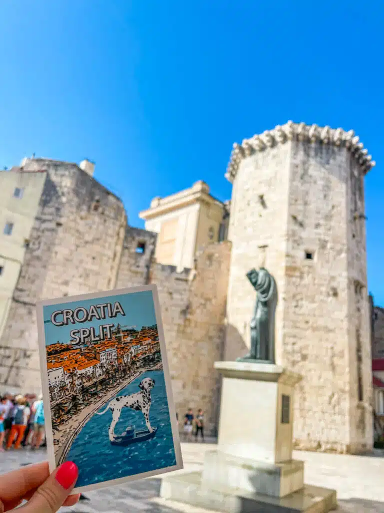 A hand holding a colorful postcard that reads Croatia Split in front of a historic stone tower and a statue in Old Town Split, Croatia, with a sunny blue sky overhead.