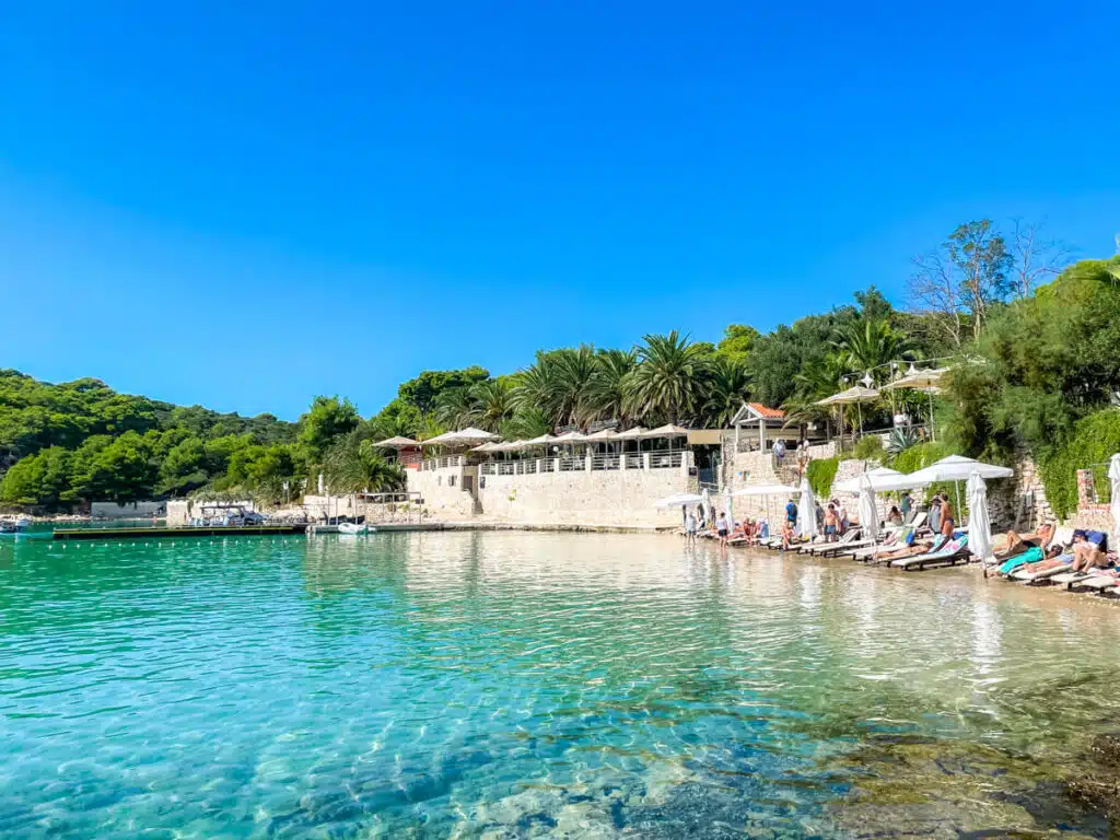 A serene beach scene with lounge chairs and umbrellas along the shore, palm trees and stone buildings behind, and vivid turquoise water in the foreground. Taken on Palmižana Beach in Croatia.