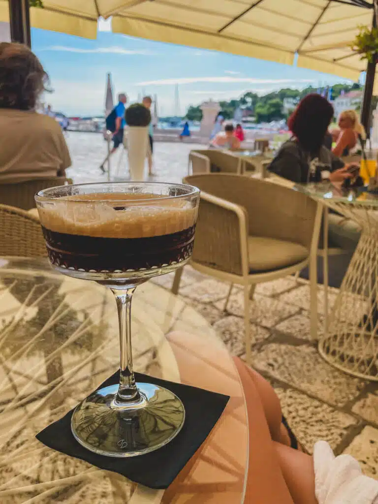 A close-up of a dark espresso martini on a glass table at a marina-side café in Hvar, with blurred tourists and yachts in the sunny background.