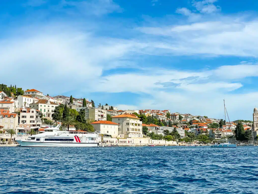 A panoramic view of Hvar in Croatia with orange rooftops and a white ferry docked along the shoreline, taken from the water. Hvar is a popular stop on a boat tour from Split.