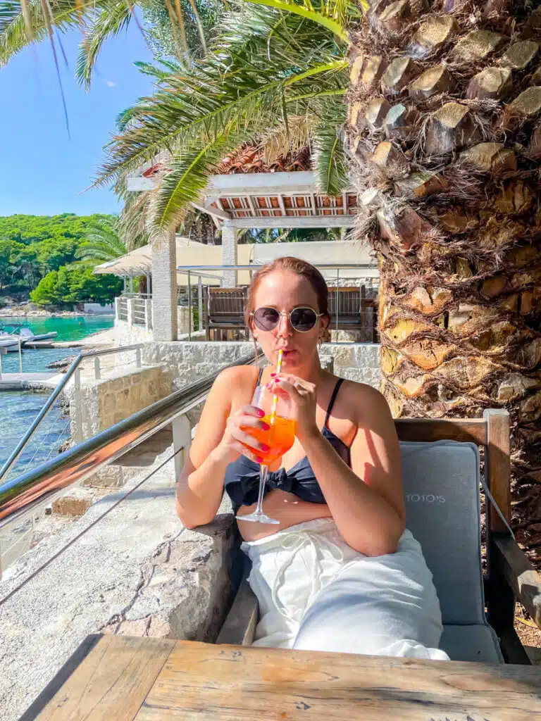 A woman relaxes at an outdoor beachside bar at Palmižana Beach in Croatia, sipping an Aperol Spritz cocktail under palm trees, dressed in white pants and a black bikini top.
