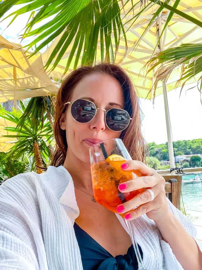 girl drinking aperol spritz in front of palm trees in pakleni islands, croatia'