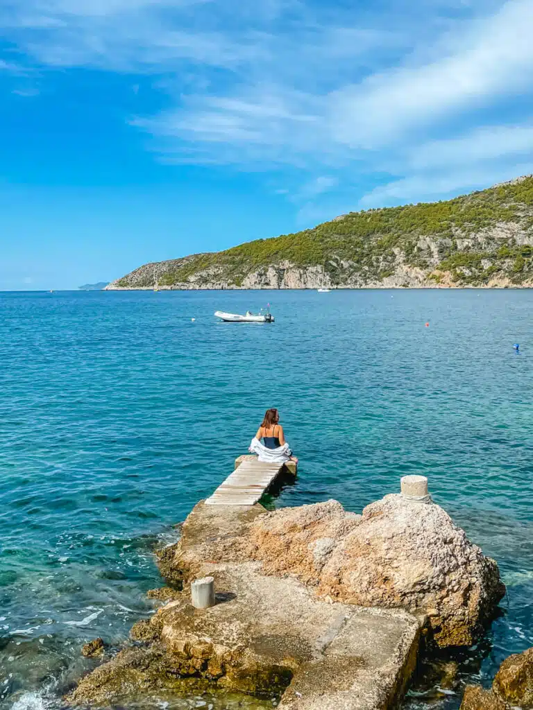 A woman sits at the end of a narrow stone pier gazing out at a calm blue sea with a single boat and green hills in the distance on the island of Vis.