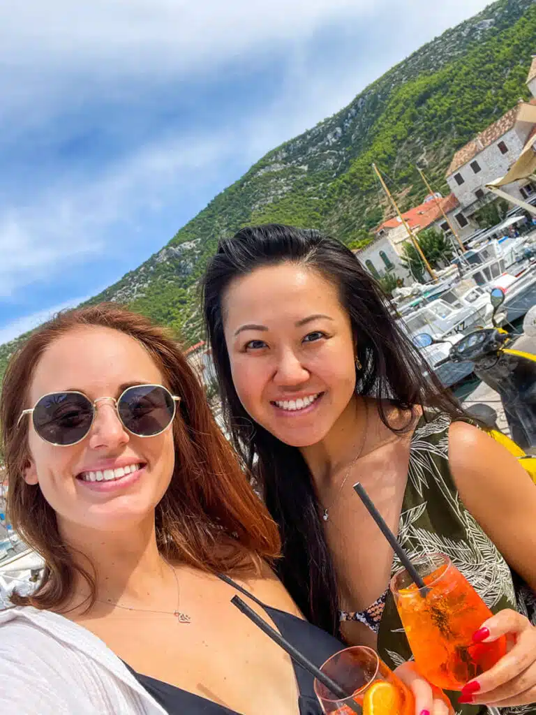 A cheerful close-up selfie of two women with Aperol Spritzes in hand, smiling in front of a marina and a lush green mountain in the town of Komiza on the island of Vis in Croatia.