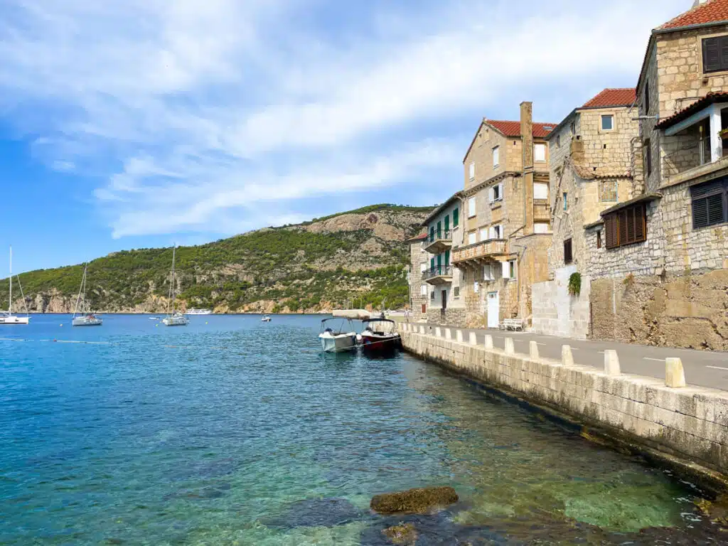 A peaceful coastal scene with calm, clear water beside a row of stone buildings and moored boats, backed by a green hillside, taken in Komiza on the island of Vis in Croatia.
