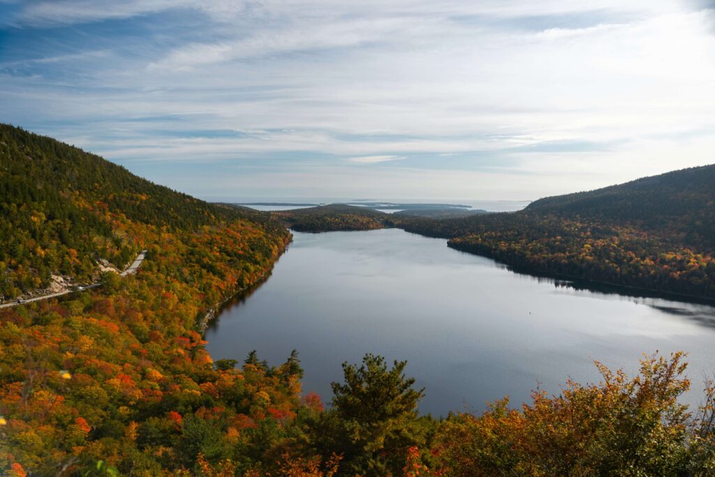 A panoramic view of Jordan Pond in Acadia National Park, flanked by forested hills showing peak fall foliage in vibrant reds, oranges, and yellows. The calm water stretches toward the horizon under a soft, cloud-streaked sky.