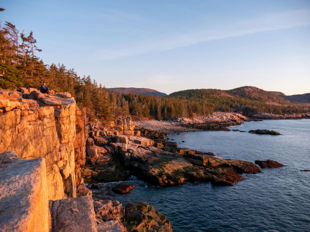 A person sits near the edge of a rugged cliff bathed in golden sunlight, overlooking the rocky coastline and forested hills of Acadia National Park in Maine. The ocean glistens below, with waves gently brushing the shoreline.