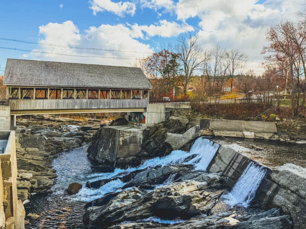 A rustic covered bridge spans a rocky river gorge with cascading waterfalls below, beside the Simon Pearce glassblowing facility in Quechee, Vermont.