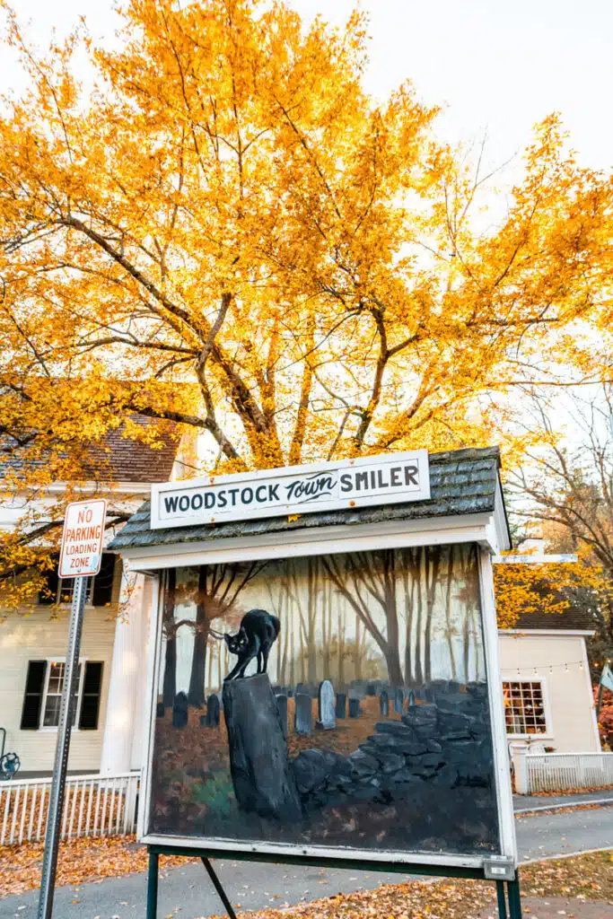 A whimsical roadside display labeled "Woodstock tOwn Smiler" featuring a painting of a black cat on a gravestone in a spooky graveyard, set against a backdrop of fiery golden leaves and a white building.