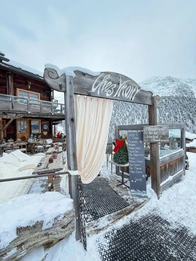 Entrance to Chez Vrony restaurant in Zermatt featuring a wooden archway with the name carved on top, cream-colored curtains, and a chalkboard drink menu beside snow-covered seating.