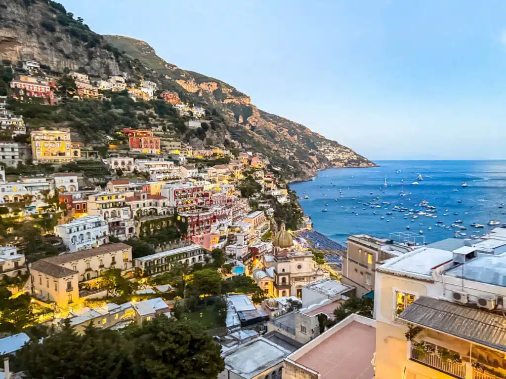 Evening view of Positano with glowing lights illuminating the hillside homes and boats anchored along the darkening sea. A dreamy twilight stop on any Amalfi Coast itinerary.
