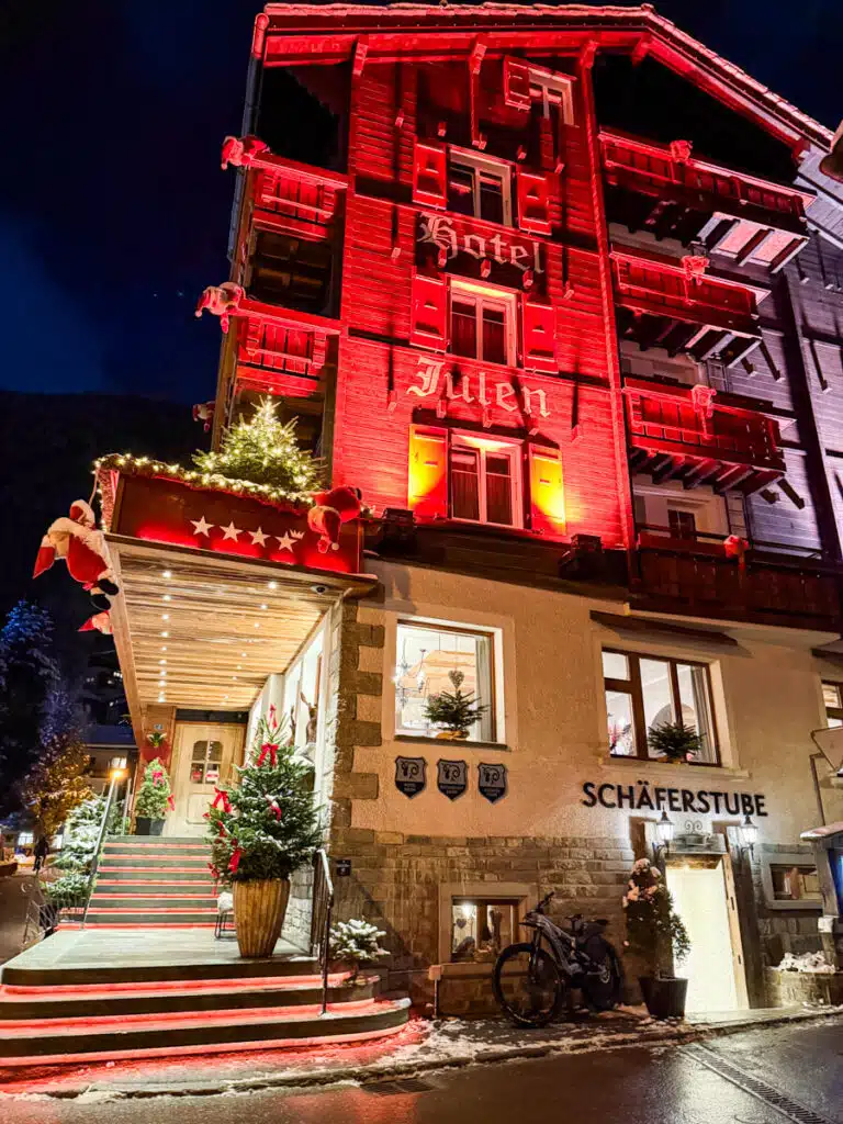 The exterior of Hotel Julen in Zermatt is lit up in red holiday lights at night, with festive decorations including Santa figurines, glowing stars, and Christmas trees leading up to the warmly lit entrance of Schäferstube restaurant.