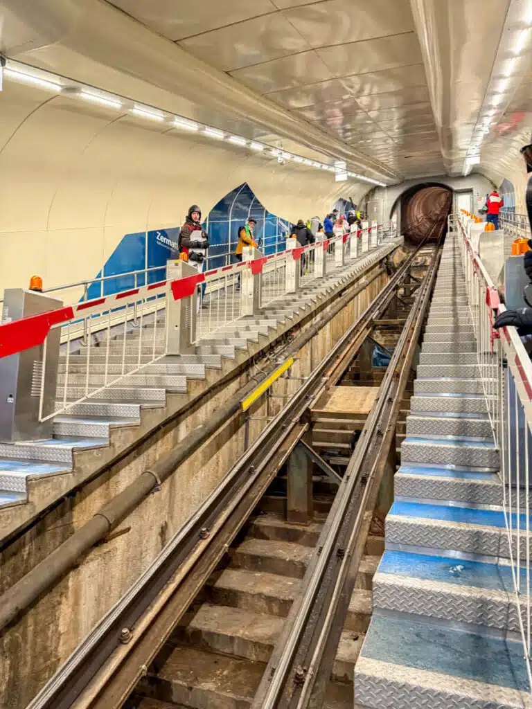 Underground funicular station in Zermatt with travelers waiting on both sides of the track as the tunnel stretches toward the mountains.