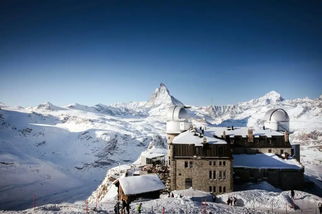 The Gornergrat Observatory sits high in the Swiss Alps, its stone building and domed telescopes surrounded by snow, with the Matterhorn rising sharply in the distance under a clear blue sky.