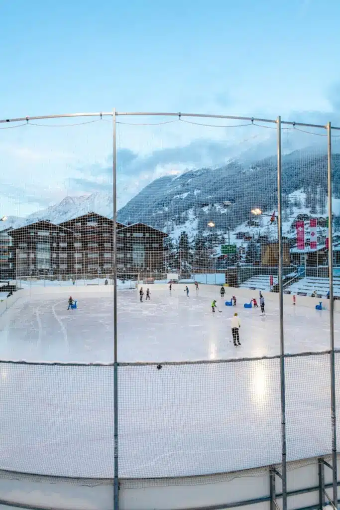 An outdoor ice rink in the snowy alpine village of Zermatt is filled with skaters and a few people playing hockey. The rink is surrounded by safety netting, with chalet-style buildings and snow-covered mountains in the background under a blue sky.