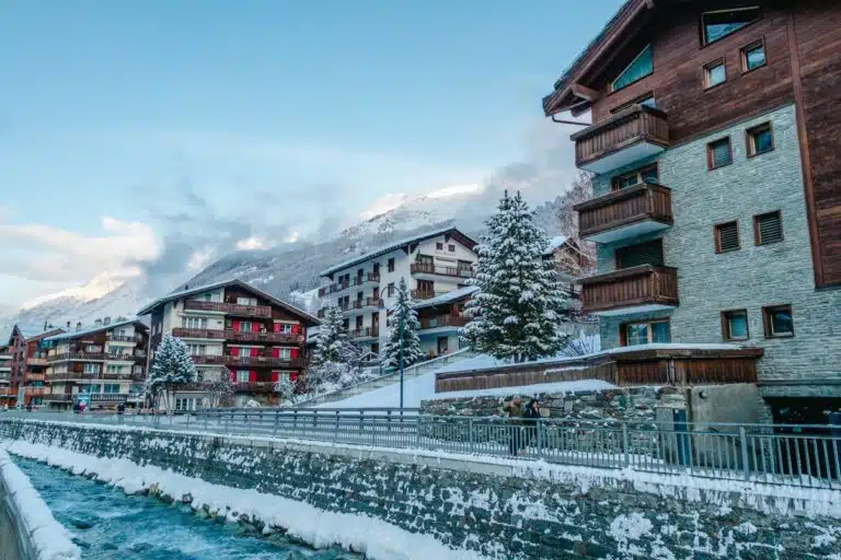 snow-frosted Zermatt with icy river, walkway, and colorful wooden hotels with blue skies
