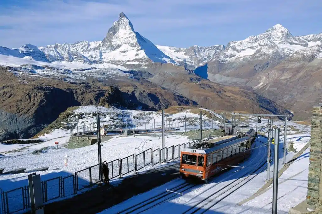 A bright red Gornergrat Bahn cogwheel train climbs the snowy mountain tracks near Zermatt, with the Matterhorn and surrounding peaks towering dramatically in the background.