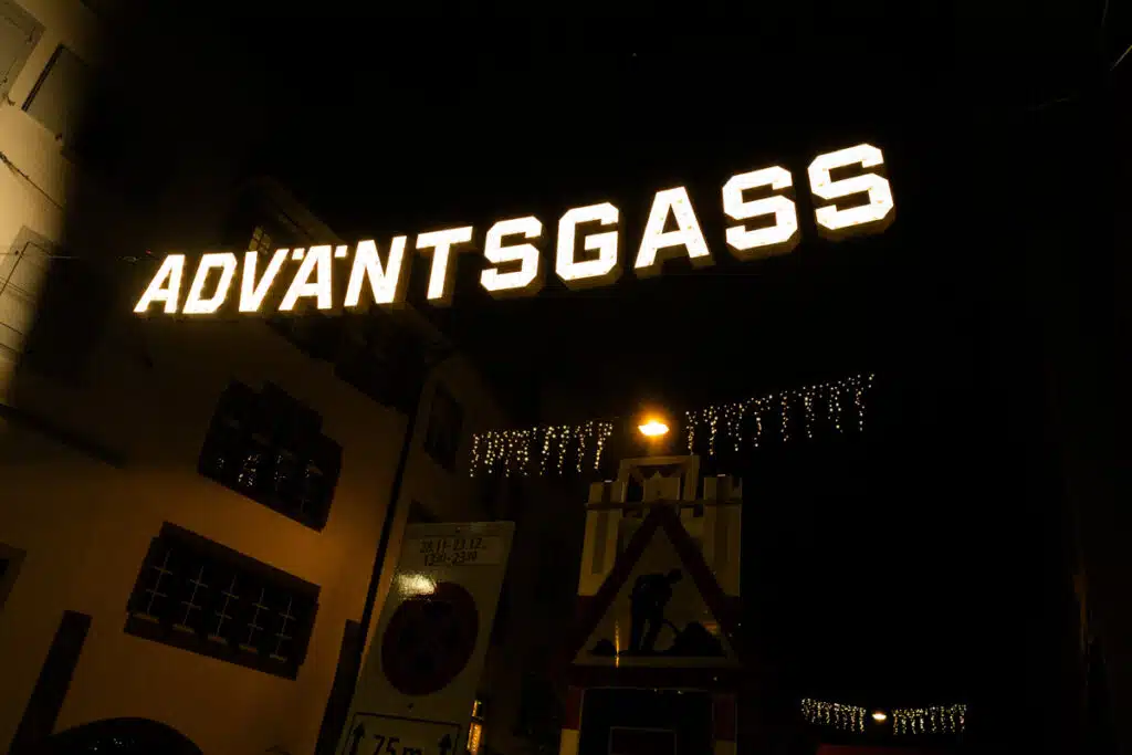 lit up sign reading "Advantsgass" hanging above dim street in Basel, Switzerland