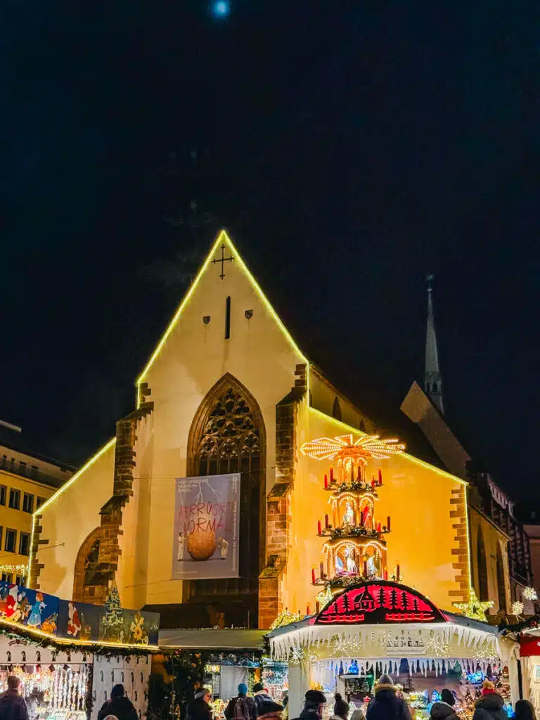A festive Christmas market at night in front of a Barfuuser church in Basel outlined with yellow lights, featuring a tall wooden pyramid decoration with red candles and a glowing nativity scene roof.