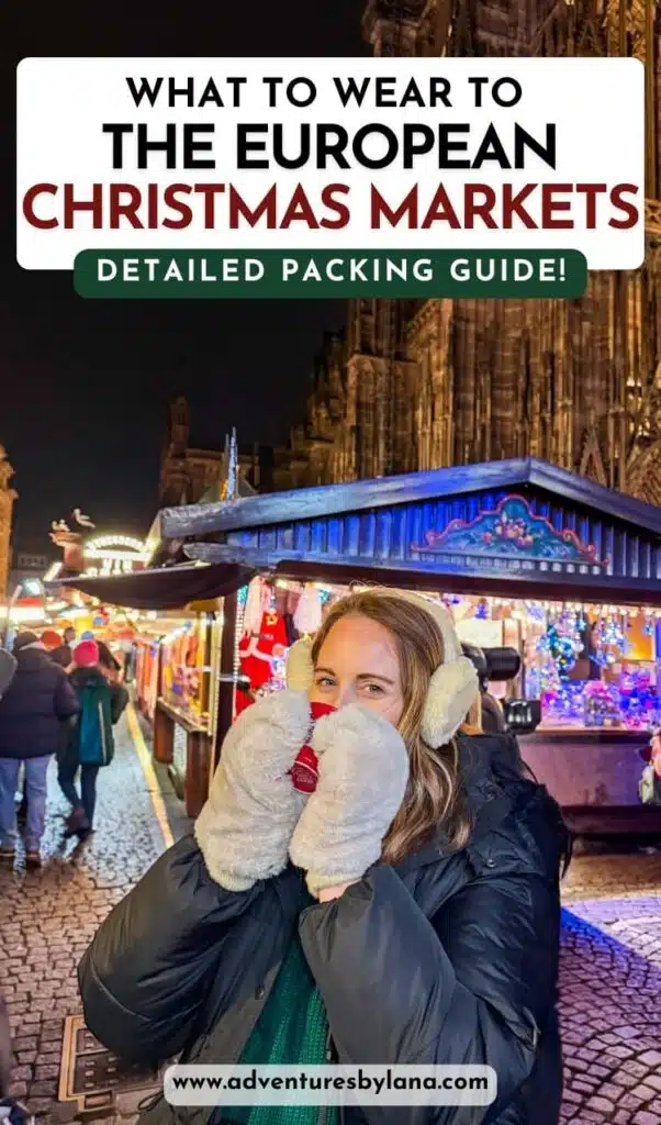A woman bundled in a black puffer coat, earmuffs, and oversized mittens holds a festive red mug at a Christmas market in Strasbourg, France. Twinkling lights, holiday decorations, and wooden stalls fill the background. Text overlay reads “What to Wear to the European Christmas Markets – Detailed Packing Guide.”
