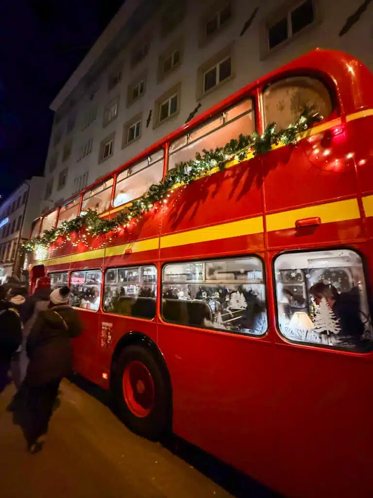 Red double-decker bus with garland and string lights, people dining inside, on Rheingasse at the Adväntsgass Christmas market in Basel