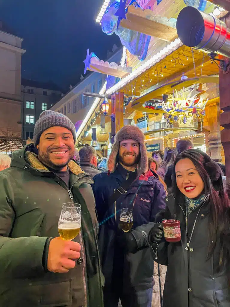 Three friends holding beers and a Christmas market mug in front of a lit-up wooden stall at the Barfüsserplatz Christmas market