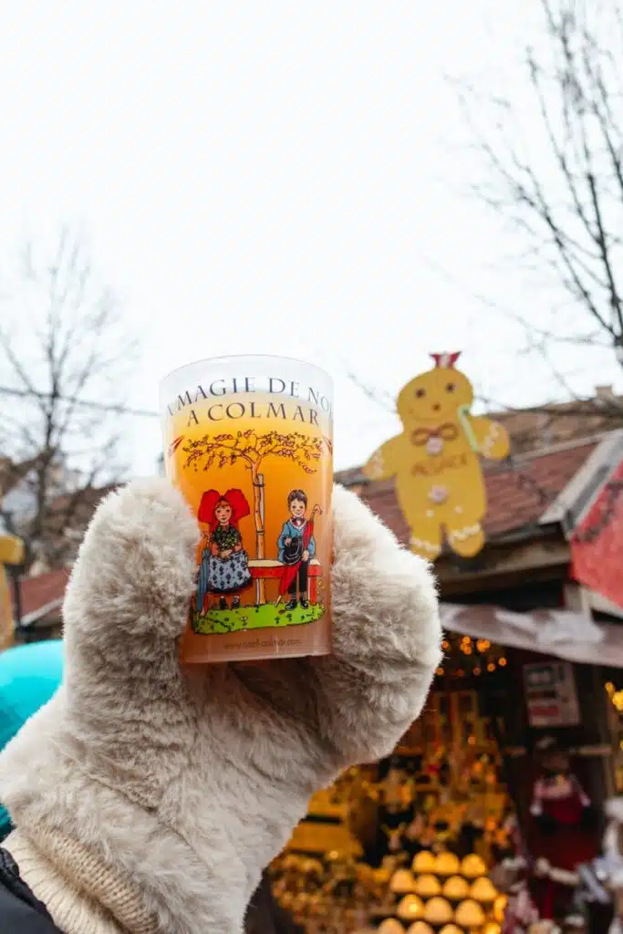 fuzzy mittened hand holding Colmar christmas cup full of hot apple cider in front of christmas stand in Colmar, France