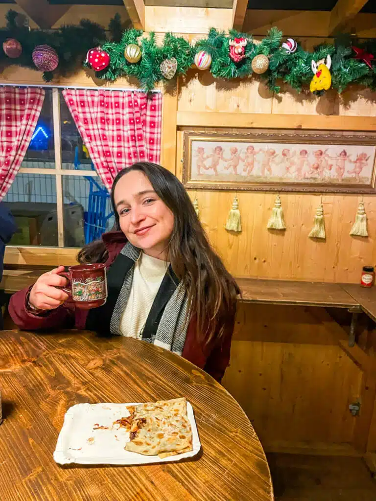 Woman holding a festive mug of glühwein next to a half-eaten crêpe inside a decorated wooden hut at the Basel Munsterplatz Christmas market