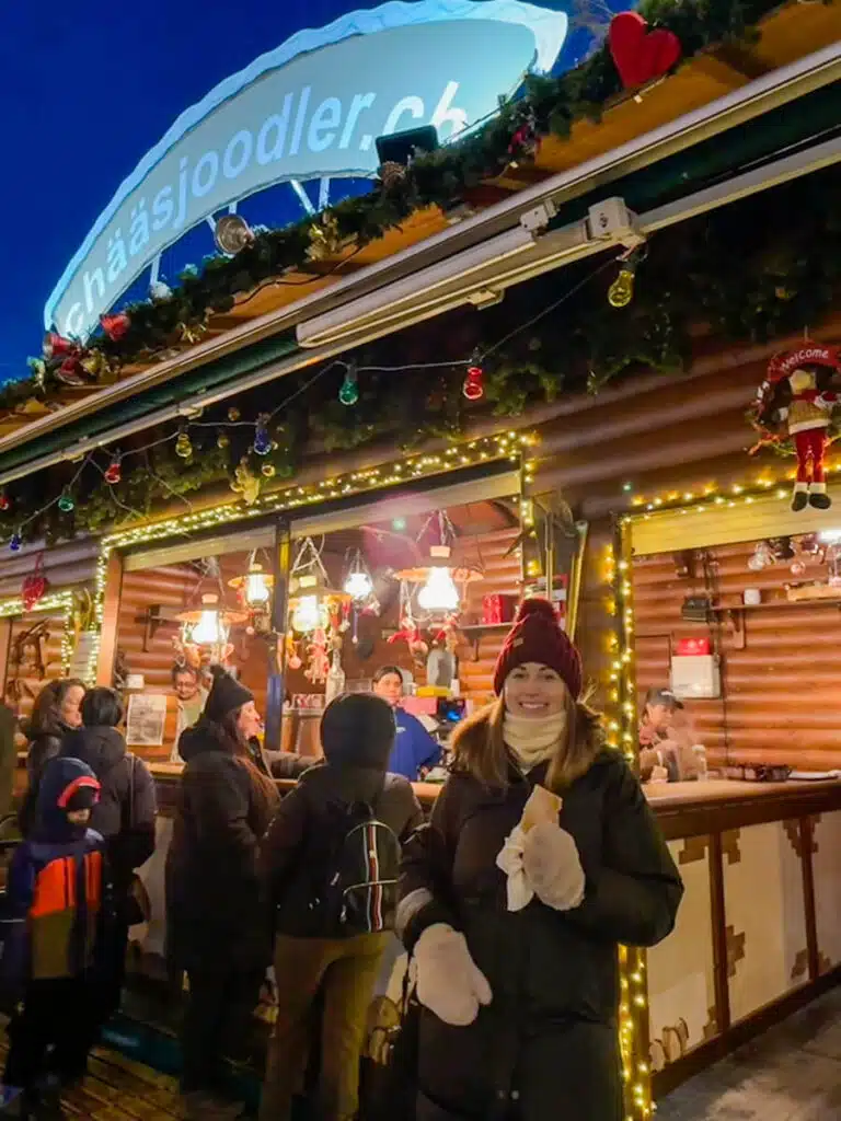 Woman in a red knit hat holding a cheese snack outside the Chääsjoodler stall at the Basel Claraplatz Christmas market at night