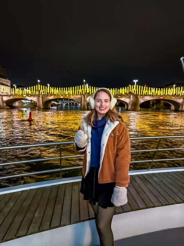 A woman in cozy winter attire, including earmuffs and mittens, smiles while holding a drink on the back of a Basel winter river cruise. Behind her, a beautifully lit bridge with yellow vertical lights reflects off the water, creating a festive and scenic atmosphere