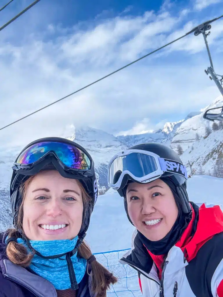 Two beginner skiers in helmets and goggles taking a selfie under a chairlift in the Sunnegga ski area, Zermatt