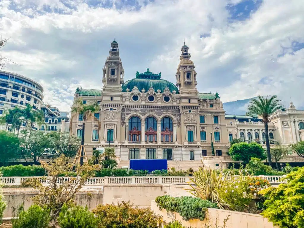 The elegant façade of the Monte Carlo Casino in Monaco is seen with green copper domes, palm trees, and manicured gardens beneath a partly cloudy sky.